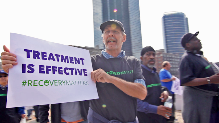 A man walks in a peaceful demonstration carrying a sign saying 'Treatment is Effective #recoverymatters.'
