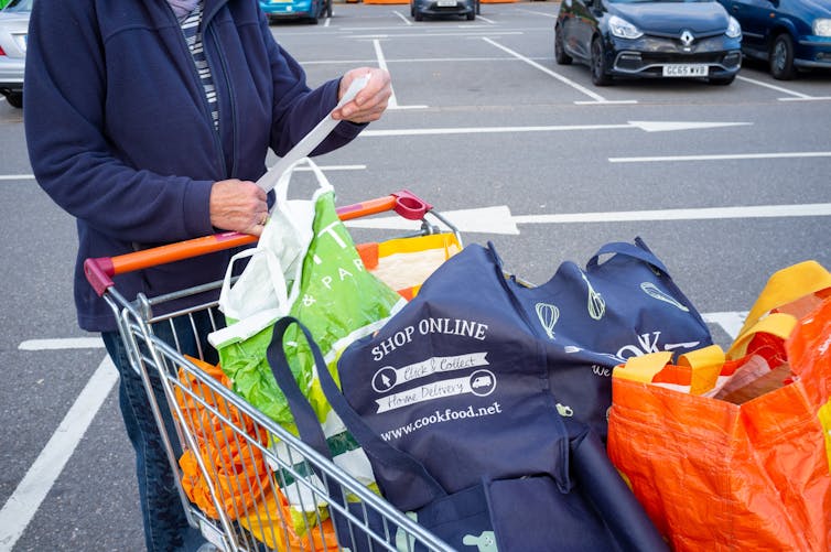 A person reads their receipt in a supermarket car park with a trolley full of shopping.