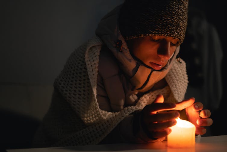 A young man wearing winter clothes indoors cups a candle in his hands.