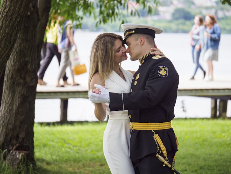 A woman in a white dress is being held by a military officer while other people walk in a park.