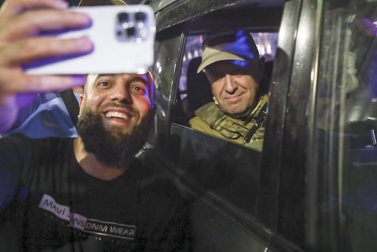 Prigozhin smiles from inside a vehicle while a bearded man takes a selfie with him