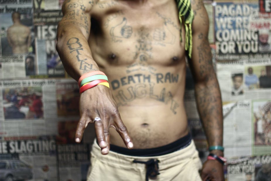 A man's torso is pictured in a room with newspaper clippings forming wallpaper. He gestures with one hand, displaying tattoos that read "outlaw" and "death raw" among many others.