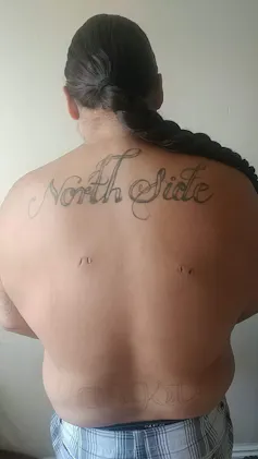 A man without a shirt pictured from behind, a prominent tattoo on his shoulders reads