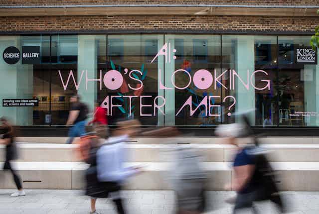 The glass outside wall of the exhibition building with the words 'AI: Who's Looking After Me' in pink lettering.