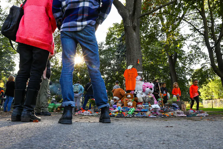 People's feet seen standing amid grass and bushes in front of a memorial showing orange shirts and stuffed animals.
