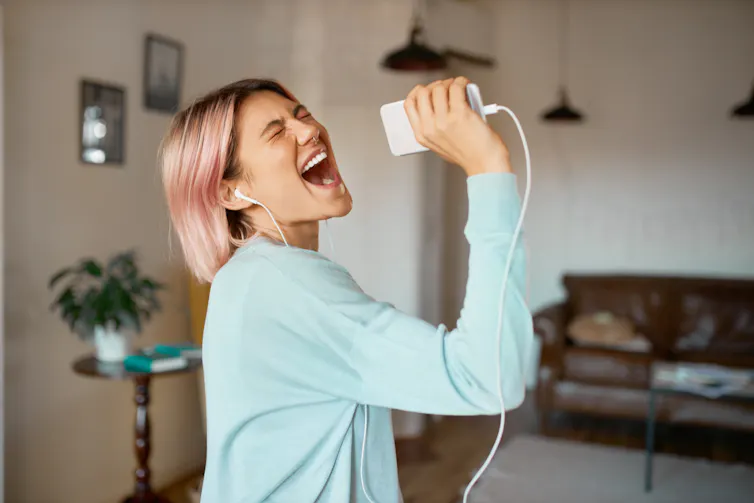 Woman with pink hair singing into music device with headphones on.