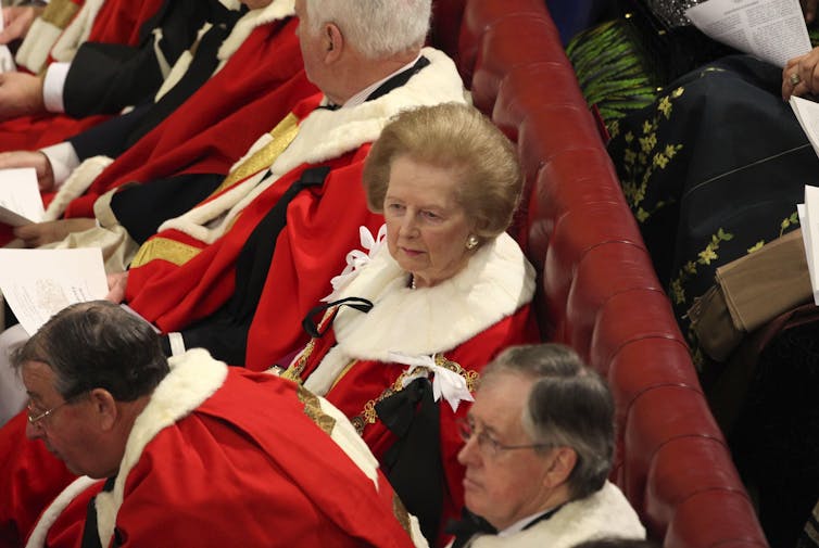 Margaret Thatcher wearing ermine, sitting on the benches of the House of Lords surrounded by other peers.