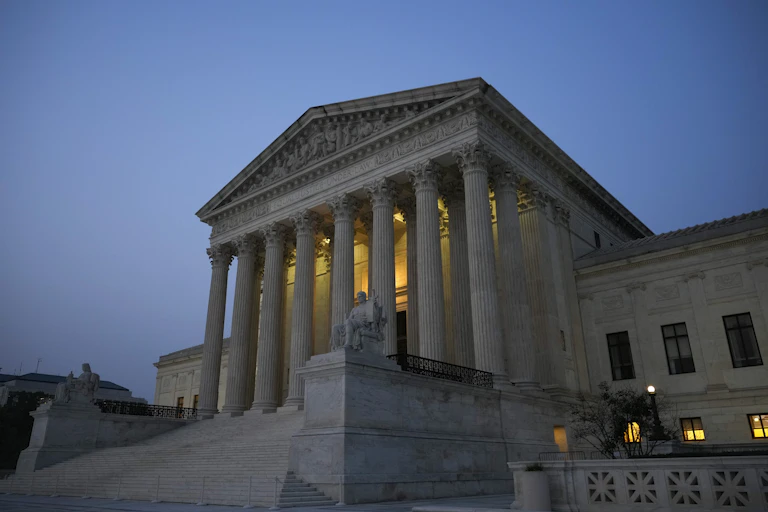 A white building with marble columns has a few lights on as the sun is going down.