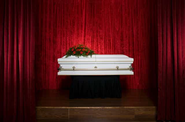 A white coffin with red flowers placed on top of it, set against a red velvet curtain.