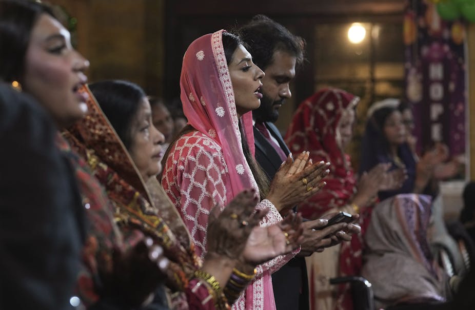Women in brightly colored dresses, heads covered, pray, alongside men, while singing and clapping.