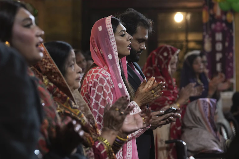 Women in brightly colored dresses, heads covered, pray, alongside men, while singing and clapping.