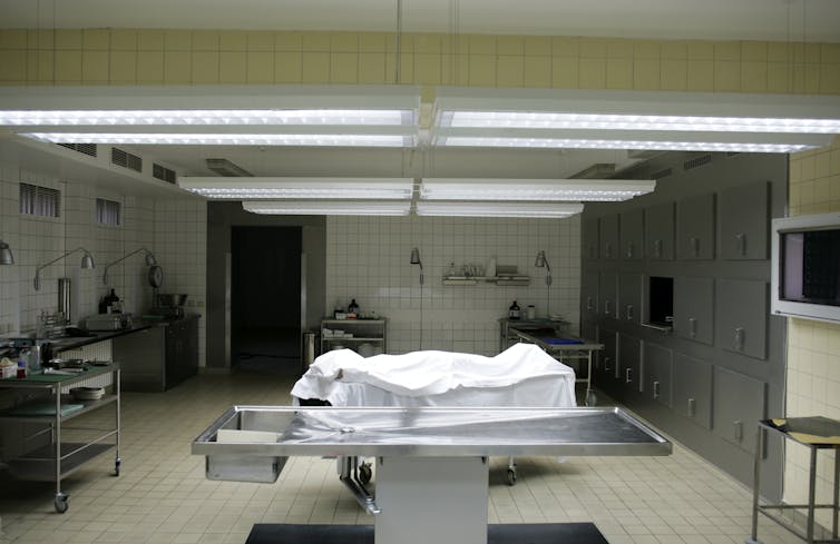 A body lies covered by a white sheet on a metal table in a white and yellow lab-like room.