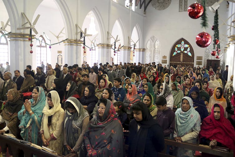 Women, with heads covered, seated in pews, inside a church with red hanging decorations.