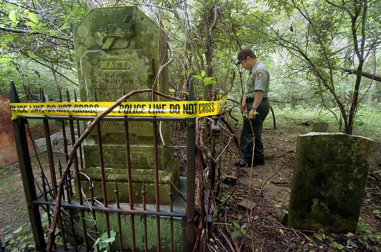 A man in a green uniform and cap walks around a tombstone with a small fence around it in a wooded area.