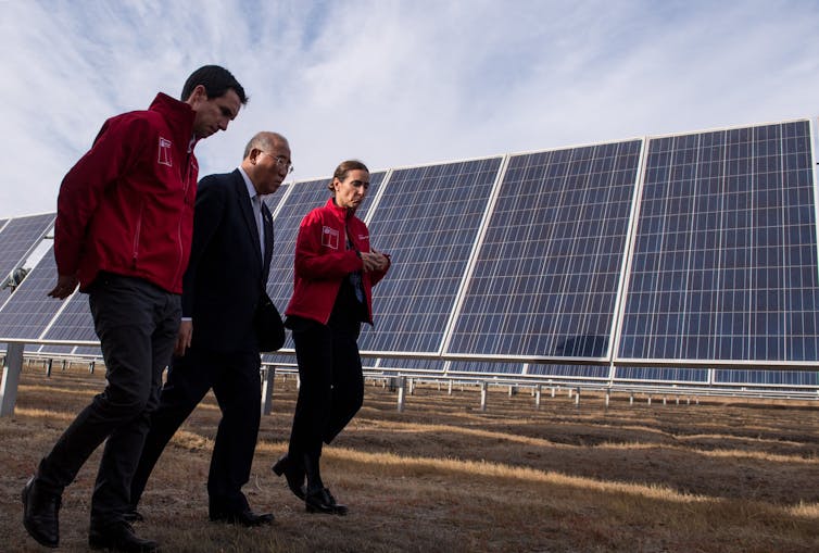 Juan Carlos Jobet and Carolina Schmidt, wearing matching fleece jackets, walk on either side Xie Zhenhua, who is wearing in a suit and tie, along a row of solar panels.