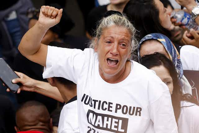 A tearful woman raising a fist amid a crowd wearing a tshir that reads 'justice for Nahel'.
