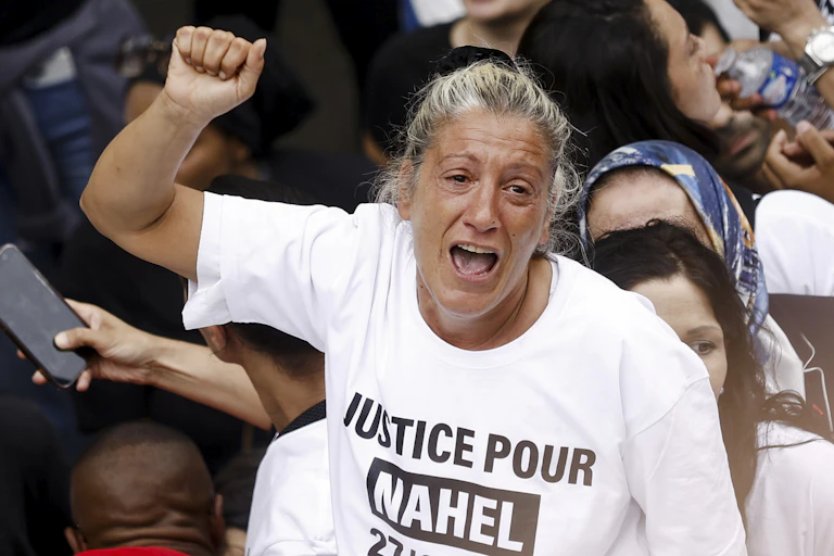 A tearful woman raising a fist amid a crowd wearing a tshir that reads 'justice for Nahel'.