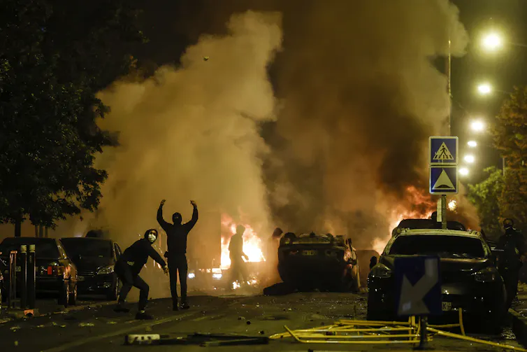 Cars burning while rioters circle in the dark on a Paris street.