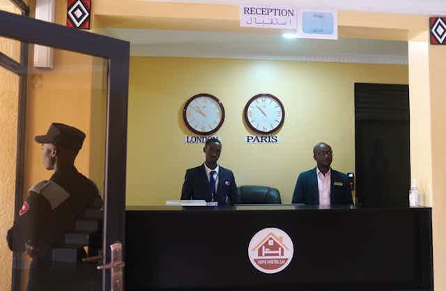 Two members of staff stand at a reception desk in front of two clocks showing the time in London and Paris. A guard in military uniform stands at the door. 