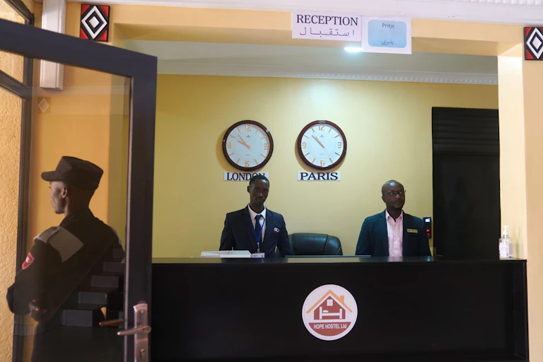 Two members of staff stand at a reception desk in front of two clocks showing the time in London and Paris. A guard in military uniform stands at the door.