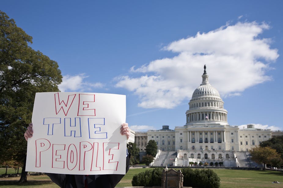 A person holds up a white sign with the words 'We the people' in front of the US Capitol building. The person's face and body is mostly obscured by the sign. 