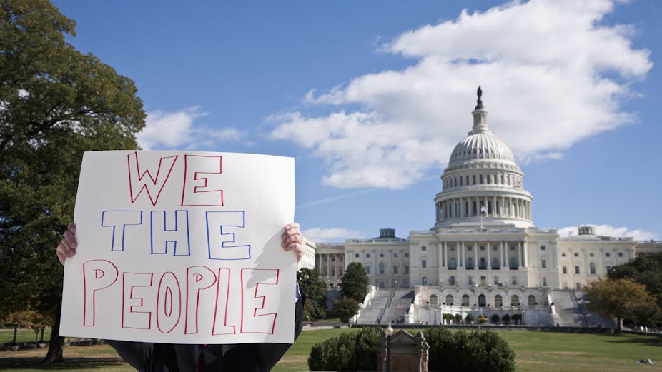 A person holds up a white sign with the words 'We the people' in front of the US Capitol building. The person's face and body is mostly obscured by the sign.