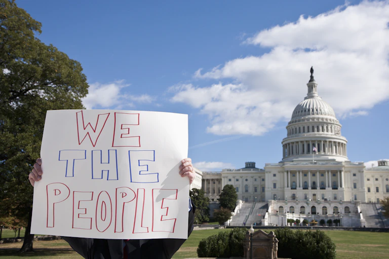 A person holds up a white sign with the words 'We the people' in front of the US Capitol building. The person's face and body is mostly obscured by the sign.