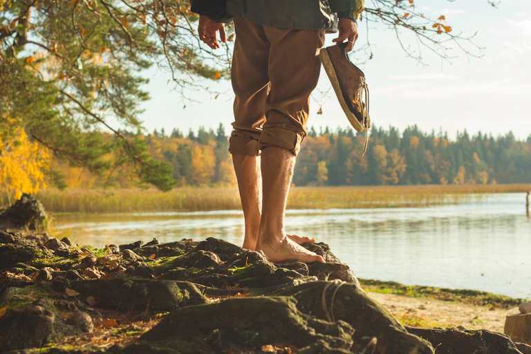 A man's legs shown stood on the bank of a river, without shoes on. He holds his trainers in his hand.
