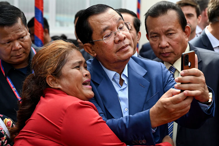 A man holds phone while onlookers behind him peer into the camera lens.