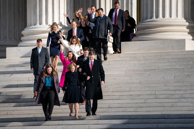 A small crowd of people in coats walk cheerfully down the steps of a building with large pillars.