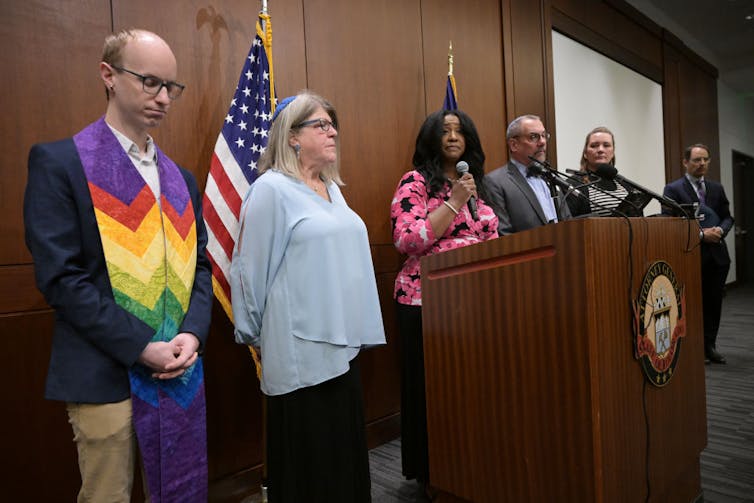 Half a dozen somber-looking people stand at the front of a room during a press conference.