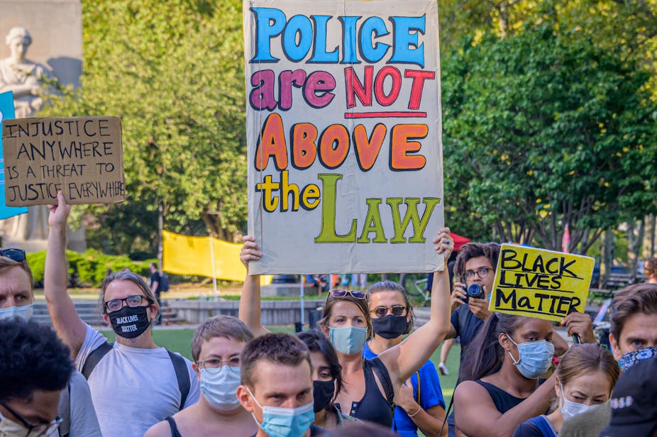 A woman wearing a face mask uses both hands to hold above her head a large, colorful, hand-written sign that reads, “Police are not above the law.” Other people, also wearing masks, stand around her.