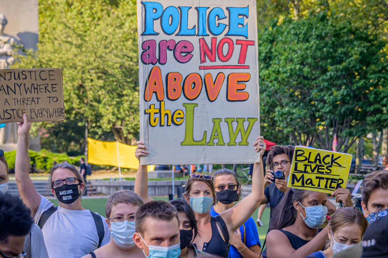 A woman wearing a face mask uses both hands to hold above her head a large, colorful, hand-written sign that reads, “Police are not above the law.” Other people, also wearing masks, stand around her.