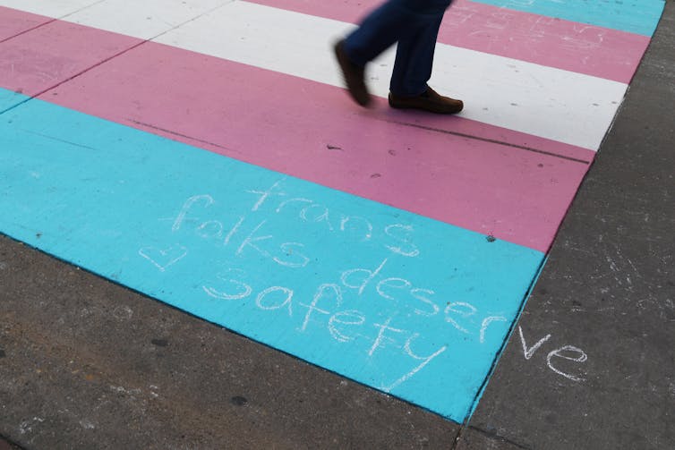 Feet seen crossing blue, pink and white colours of a trans crosswalk on a road.