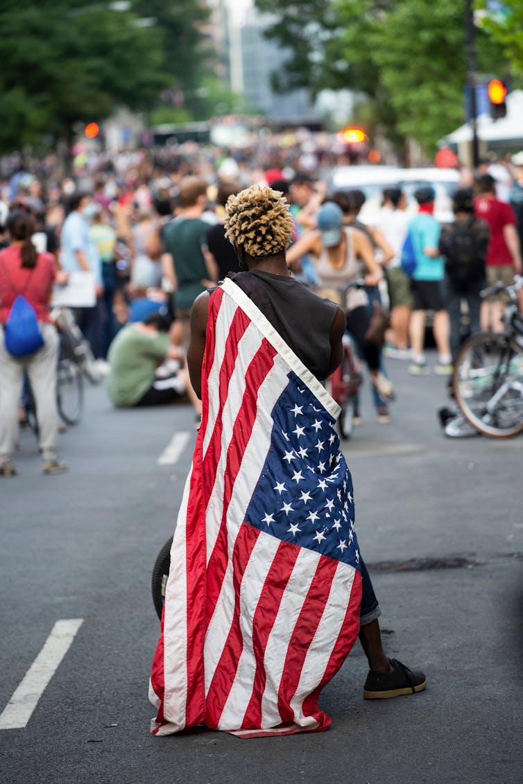 A man draped in the US flag, with his back to the camera, sits on a motorized bicycle. A crowd of people stand yards away from him with their backs to the camera as well.