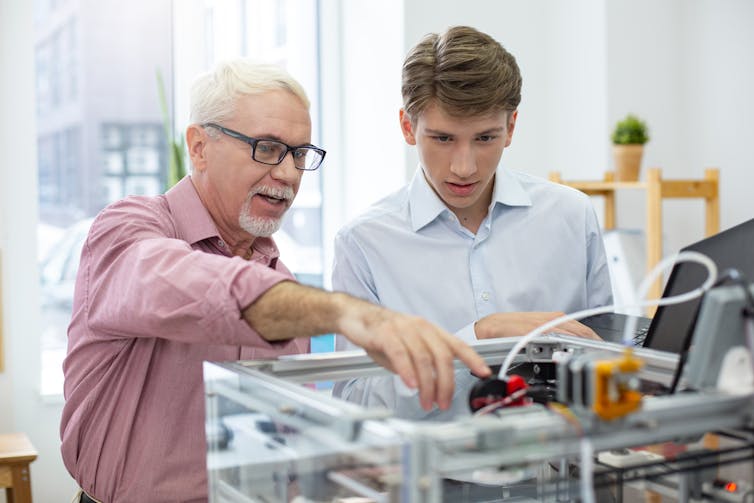 Older employer shows young male trainee how a piece of electrical equipment works.