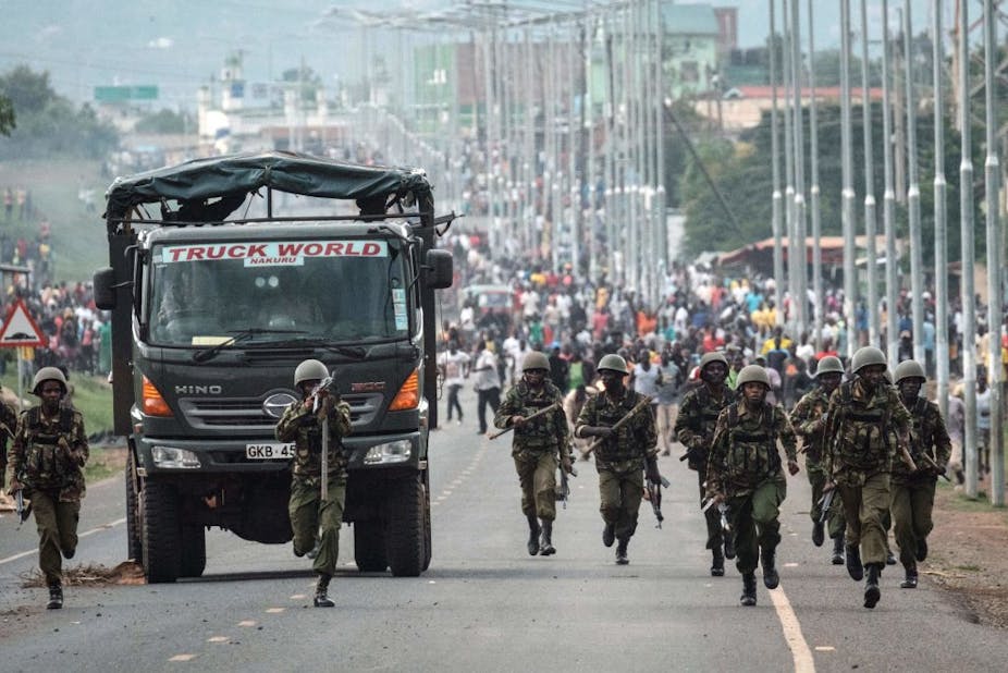 Men wearing military fatigues and helmets and holding teargas guns run ahead of a crowd of civilians, with a military truck keeping pace with the officers.