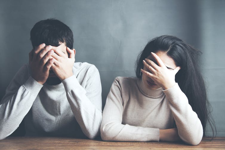 A young, heterosexual couple with dark hair, sitting side by side, both covering their faces with their hands