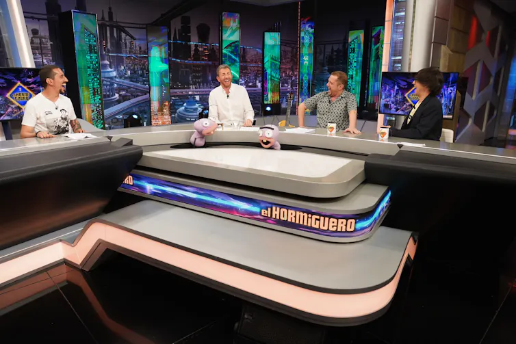Cuatro hombres ríen sentados en una mesa en forma de U.