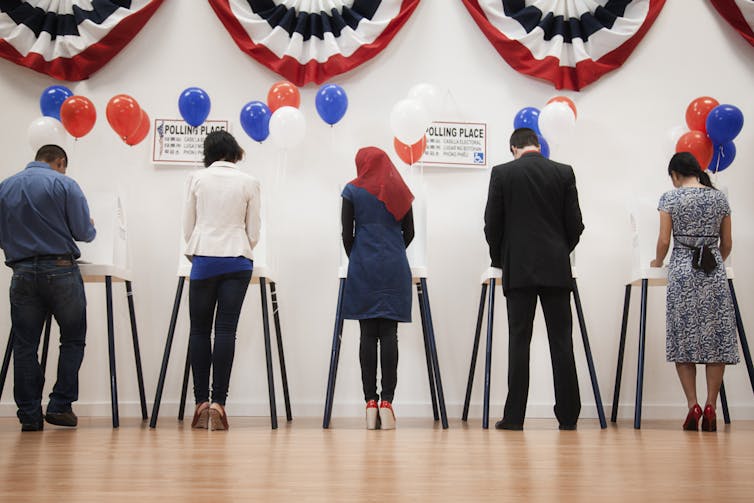 Five people with their backs to the camera vote at small booths in a room with bunting in the colors of the American flag.