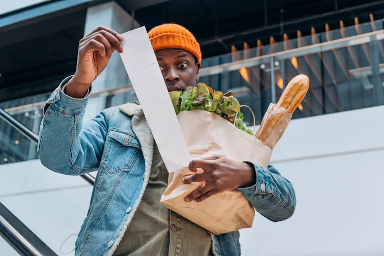 Man with orange hat and paper bag of groceries, looking at a long receipt, eyes wide.