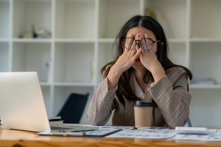 A young woman sitting at an office desk, looking very tired and rubbing her eyes behind her glasses