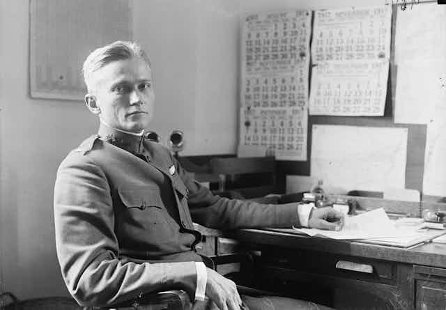 Un homme blond en uniforme regarde la caméra, assis devant un bureau rempli de papiers.