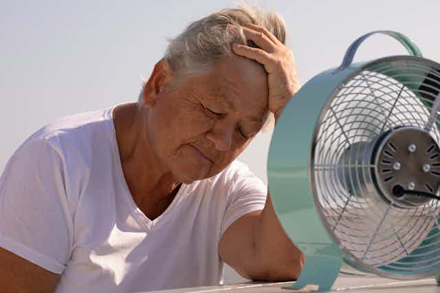 An older woman a T-shirt leans on her elbow toward a small electric fan on a table top. 