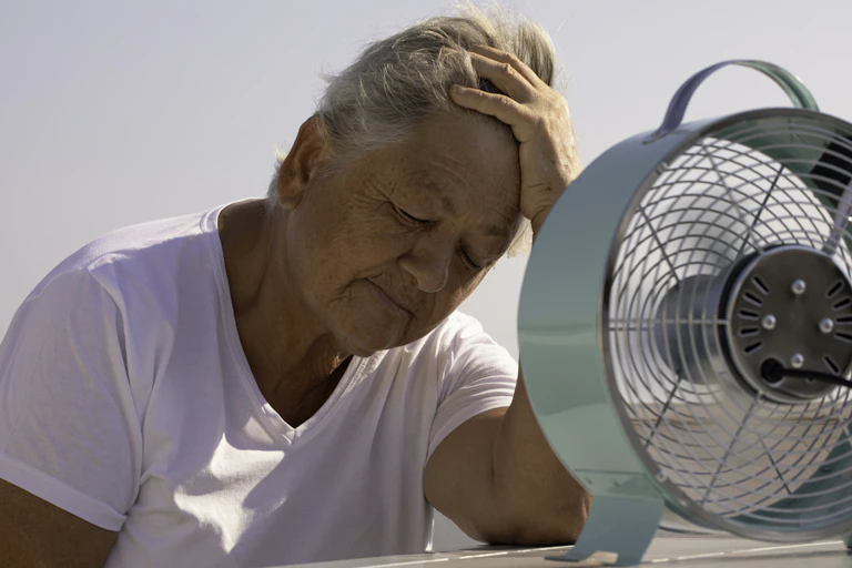 An older woman a T-shirt leans on her elbow toward a small electric fan on a table top.