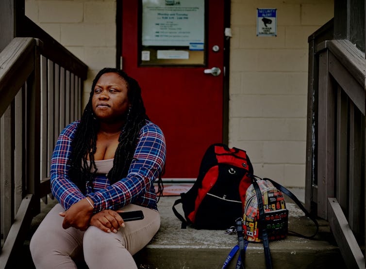 A woman sits on wooden steps outside a door. Two backpacks, one belonging to a small child, sit on the steps beside her.