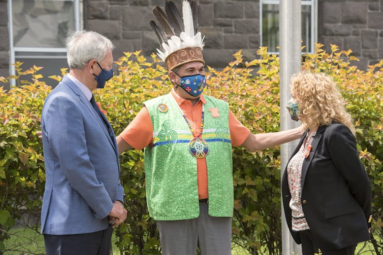 A man and a woman in masks stand on either side of an Indigenous chief, also in a mask.