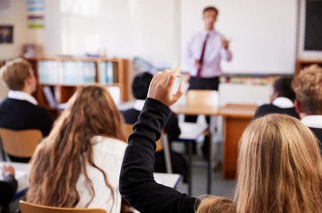 A student raises her hand in class, as teacher stands at the front.