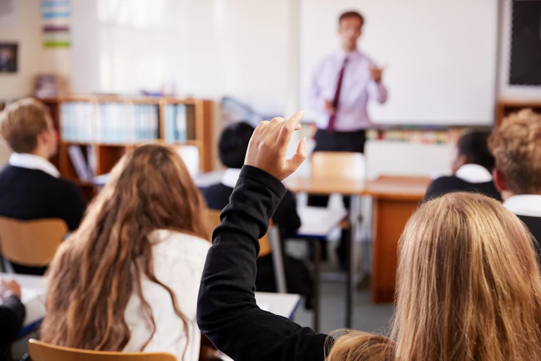 A student raises her hand in class, as teacher stands at the front.