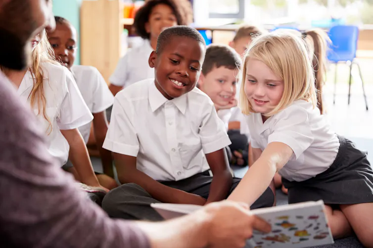 A teachers reads to young students, who are sitting on the ground.